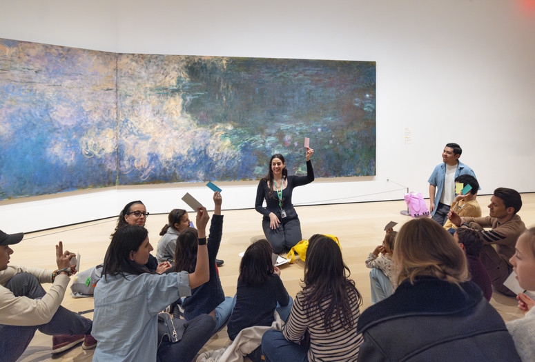 An educator kneels in front of an artwork, holding a pink color card, and leads a discussion about the art with a group of families. Photo: On White Wall, Inc.
