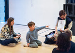 In this image from an Arte en Familia/Art Among Family event, a mother looks on as a child points to an educator holding a piece of paper in front of a sculpture. Photo: On White Wall, Inc. Digital Image © 2025 The Museum of Modern Art, New York