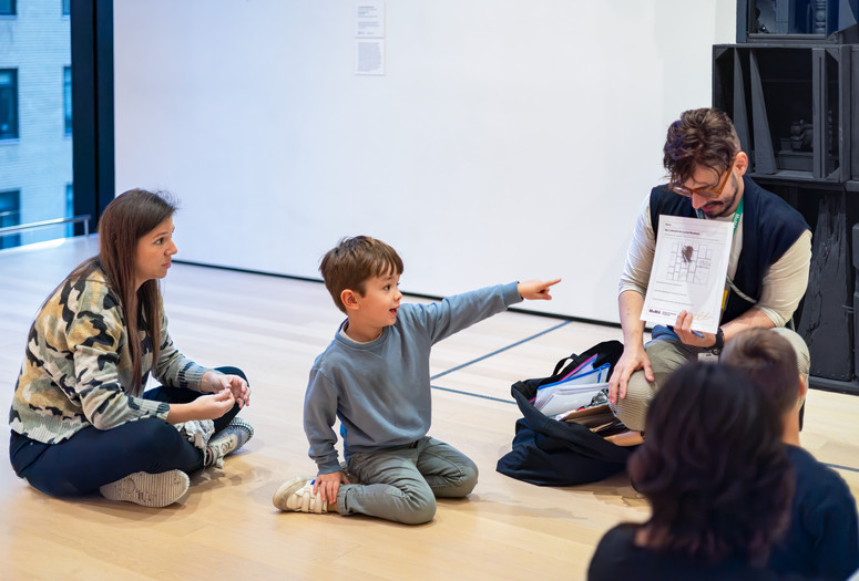 In this image from an Arte en Familia/Art Among Family event, a mother looks on as a child points to an educator holding a piece of paper in front of a sculpture. Photo: On White Wall, Inc. Digital Image © 2025 The Museum of Modern Art, New York