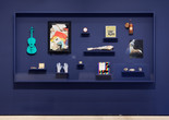 A display case with a dark blue background contains various objects, including a turquoise violin, gloves, and books. Items are arranged on black shelves, contrasting against the backdrop. Installation view, The Art of Assemblage, The Museum of Modern Art, New York, March 28, 2025–ongoing. Photographed in April 2025. Photo: Martin Parsekian. Digital Image © 2025 The Museum of Modern Art, New York