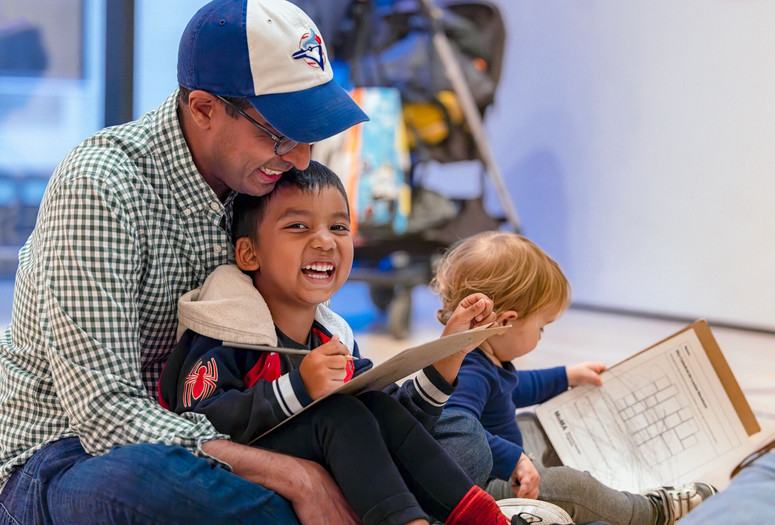 A father in a baseball cap looks down at his laughing child, who is holding a pencil and has drawing materials in his lap. This photo was taken at the event Arte en Familia/Art Among Family on October 26, 2024. Photo: On White Wall. Digital Image © 2025 The Museum of Modern Art, New York