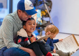 A father in a baseball cap looks down at his laughing child, who is holding a pencil and has drawing materials in his lap. This photo was taken at the event Arte en Familia/Art Among Family on October 26, 2024. Photo: On White Wall. Digital Image © 2025 The Museum of Modern Art, New York
