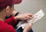A child holds their composition of brown string on a white sticky board Photo: On White Wall Inc.