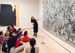 A group of people sit on a gallery floor as an educator gestures toward a large abstract painting with black, white, and beige splatters. Another bold painting with dark tones hangs nearby. Photo: On White Wall