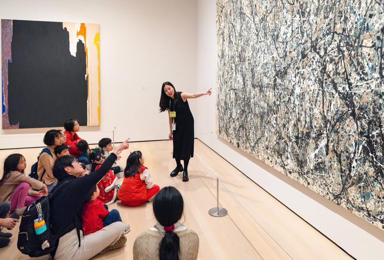 A group of people sit on a gallery floor as an educator gestures toward a large abstract painting with black, white, and beige splatters. Another bold painting with dark tones hangs nearby. Photo: On White Wall