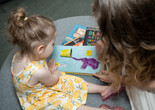 A child and an adult look at a picture book with an elephant. There are other books piled underneath, and they are sitting on a circular mat. Photo: Austin Donohue