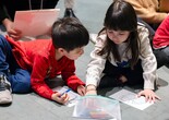 Children in casual clothes sit on the floor, drawing with pastels on paper. The scene features red and white clothing, with a clear pencil case nearby. Photo: On White Wall Inc.