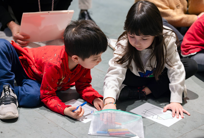 Children in casual clothes sit on the floor, drawing with pastels on paper. The scene features red and white clothing, with a clear pencil case nearby. Photo: On White Wall Inc.