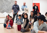 Children hold up sketches of yarn in a gallery with a large monochrome artwork on the wall. The setting features bright, colorful clothing and a mix of seated and standing individuals. Photo: On White Wall Inc.