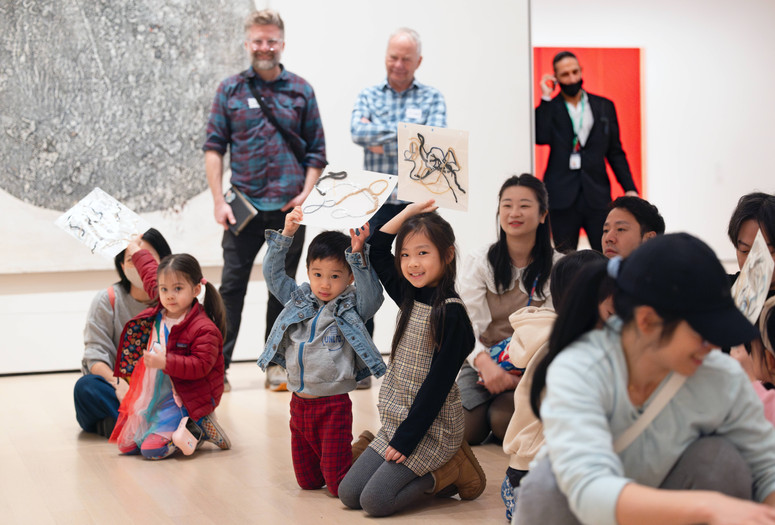 Children hold up sketches of yarn in a gallery with a large monochrome artwork on the wall. The setting features bright, colorful clothing and a mix of seated and standing individuals. Photo: On White Wall Inc.