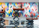 Installation view of the exhibition Odili Donald Odita: Songs from Life, The Museum of Modern Art, New York, April 8, 2025–Spring 2026. Photo: Jonathan Dorado. Image description: Three people stand on three separate scissor lifts as they paint a mural of colorful shapes on a white wall.