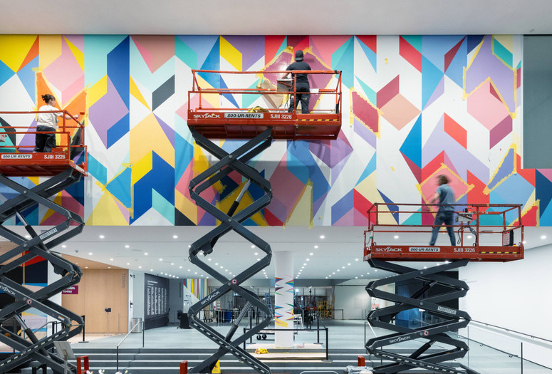 Installation view of the exhibition Odili Donald Odita: Songs from Life, The Museum of Modern Art, New York, April 8, 2025–Spring 2026. Photo: Jonathan Dorado. Image description: Three people stand on three separate scissor lifts as they paint a mural of colorful shapes on a white wall.