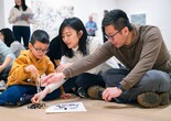 A group of people sits on the floor, engaging with a jumble of yarn on a paper. The scene is vibrant, highlighting beige and blue tones. Photo: On White Wall