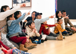 A group of adults and children sit on a wooden floor, pointing and looking in the same direction. They are wearing casual clothing in shades of blue, red, and beige. Photo: On White Wall Inc.