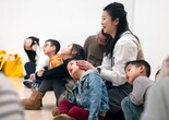 An adult holds their child’s head and turns it to the side as they sit among a group of families during a gallery talk. Photo: On White Wall, Inc.