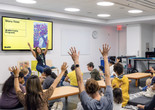 A group of families sit on the floor in a classroom raising their arms as an educator leads them through an activity Photo: On White Wall, Inc.
