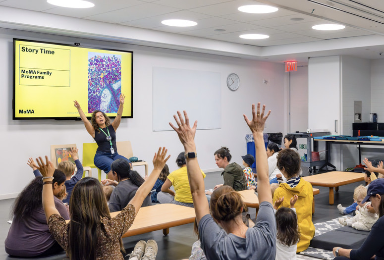 A group of families sit on the floor in a classroom raising their arms as an educator leads them through an activity Photo: On White Wall, Inc.