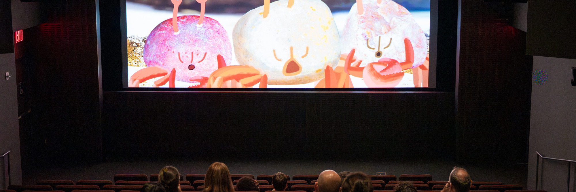 Crab Story. 2017. Russia/Australia. Directed by Tatiana Poliektova, Filippo Rivetti. What happens when a crab finds a pineapple on a beach? 4 min. Photo credit: On White Wall Inc. Alt text: A group of families is shown from behind, sitting in a movie theater and watching an animated film. On the screen, three animated crabs are featured.