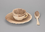 A teacup, saucer, and spoon covered in light brown fur are set against a plain grey background. The fur is thick and evenly distributed. Meret Oppenheim. Object. 1936. Fur-covered cup, saucer, and spoon. The Museum of Modern Art, New York. Purchase. © 2025 Artists Rights Society (ARS), New York/Pro Litteris, Zurich