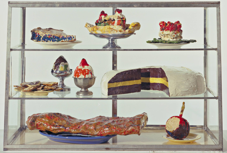 Claes Oldenburg. Pastry Case, I. 1961–62. Burlap and muslin soaked in plaster, painted with enamel, metal bowls, and ceramic plates in glass-and-metal case. The Museum of Modern Art, New York. The Sidney and Harriet Janis Collection. © 2025 Claes Oldenburg Image description: A display case features painted plaster sculptures of desserts on silver stands and plates. Predominant colors are red, white, yellow, brown, and blue.