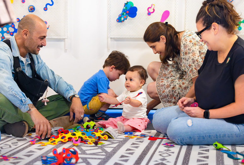 Young children and adults sit on a patterned mat, looking at one another and playing with colorful shapes. Photo: On White Wall, Inc.