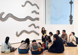 A group of children and adults and a teaching artist sit in front of an artwork. Artwork: Rosemarie Castoro. Side by Side. 1972. Modeling paste, gesso, and graphite on board, five pieces. The Museum of Modern Art, New York. Purchase. © The Estate of Rosemarie Castoro. Photo: On White Wall, Inc.