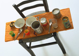 A mixed media sculpture featuring a black chair with a wooden seat covered in dishes, cans, and utensils. The predominant colors are orange, silver, and black. Shown: Daniel Spoerri. Kichka’s Breakfast I. 1960. Wood chair hung on wall with board across seat, coffeepot, tumbler, china, eggcups, eggshells, cigarette butts, spoons, tin cans, and other materials. The Museum of Modern Art, New York. Philip Johnson Fund. © 2026 Daniel Spoerri/Artists Rights Society (ARS), New York/ProLitteris, Switzerland