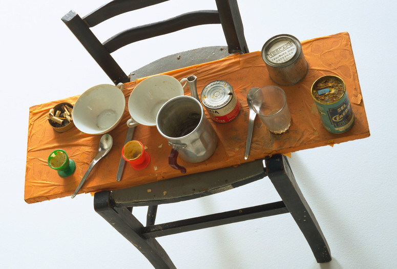 A mixed media sculpture featuring a black chair with a wooden seat covered in dishes, cans, and utensils. The predominant colors are orange, silver, and black. Shown: Daniel Spoerri. Kichka’s Breakfast I. 1960. Wood chair hung on wall with board across seat, coffeepot, tumbler, china, eggcups, eggshells, cigarette butts, spoons, tin cans, and other materials. The Museum of Modern Art, New York. Philip Johnson Fund. © 2026 Daniel Spoerri/Artists Rights Society (ARS), New York/ProLitteris, Switzerland
