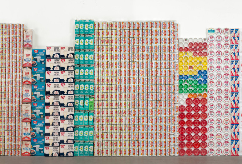 An artwork made with stacked empty containers and packaging is displayed in a gallery. Shown: George Maciunas. One Year. 1973–74. Various empty containers and packaging. Publisher: Fluxus. The Museum of Modern Art, New York. The Gilbert and Lila Silverman Fluxus Collection Gift. © 2026 George Maciunas. Used by permission