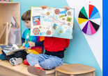 Two children sit on bench reading picture books. One holds the book on their lap, and one holds the book with the pages facing out toward the camera. Photo: On White Wall, Inc.