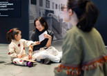 In the background, a parent and child sit on the floor and look at each other. In the foreground the back of an educator is shown. Photo: On White Wall, Inc.