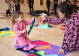 A child dressed in pink holds up a blue felt shape while she looks up at a MoMA Learning Specialist in a floral shirt. Photo: Alycia Kravitz Photography, Inc. © 2026 The Museum of Modern Art, New York
