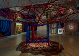 Installation view of Frida and Diego: The Last Dream, The Museum of Modern Art, New York, March 21–September 12, 2026. Photo: Jonathan Dorado. Digital image © The Museum of Modern Art, New York Image description: An installation view of a dimly lit exhibition gallery. A blood-red-colored tree in the center of the room is rooted to a mound of clay-colored earth. The tree grows up through a vibrant blue four-poster bed. Its gnarled branches reach up to cradle a large mirror hanging from the ceiling. The mirror reflects the tree within the bed below it. Paintings by the artists Frida Kahlo and Diego Rivera are spotlit on the walls in the background, framed by pulled-back blue theater curtains.