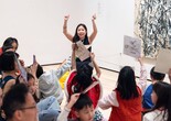 An educator holds up both arms in front of a group of families with children who hold up their yarn collages. Photo: On White Wall, Inc.