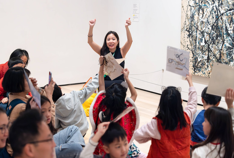 An educator holds up both arms in front of a group of families with children who hold up their yarn collages. Photo: On White Wall, Inc.