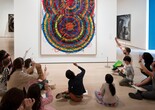 An educator leads a discussion in front of a colorful artwork by Atsuko Tanaka featuring curved lines and squiggles. Families hold up pencils in the air to trace the lines in the artwork. Photo: Martin Seck