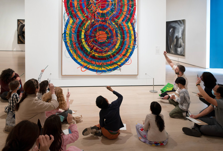 An educator leads a discussion in front of a colorful artwork by Atsuko Tanaka featuring curved lines and squiggles. Families hold up pencils in the air to trace the lines in the artwork. Photo: Martin Seck