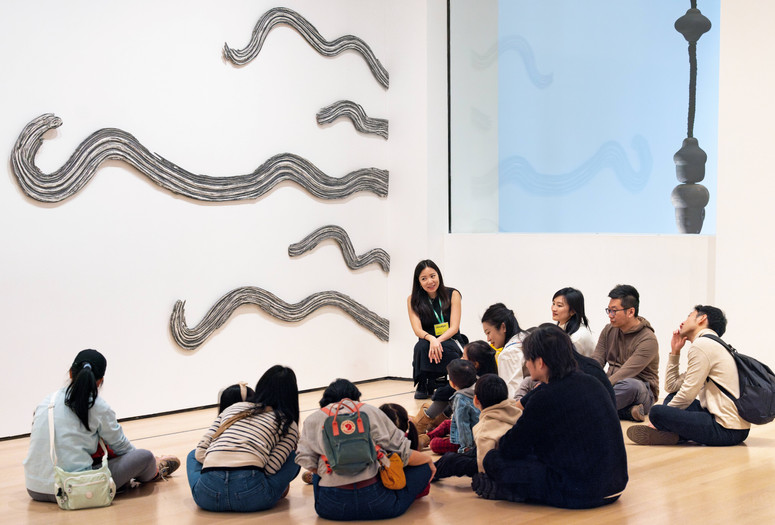 A group of children and adults and a teaching artist sit in front of the artwork Side by Side (1972) by Rosemarie Castoro. Photo: On White Wall, Inc.