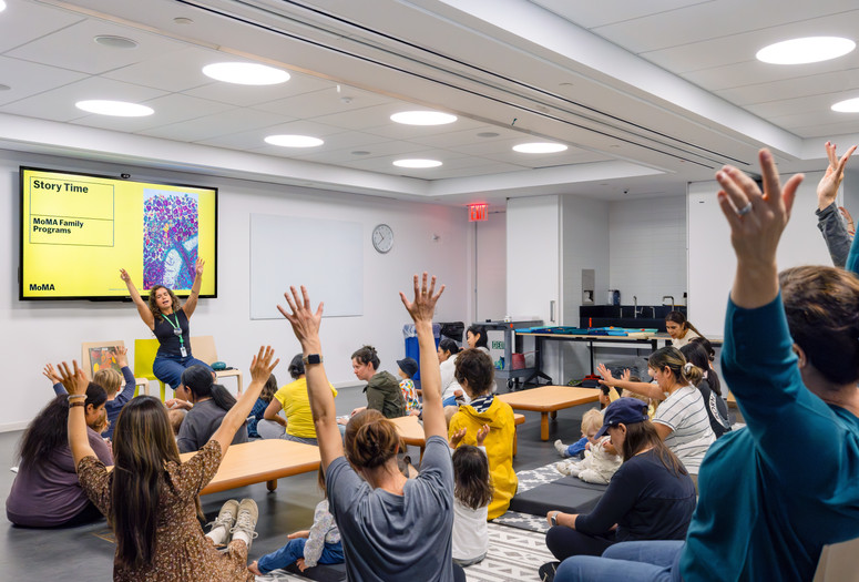 An educator sits in front of families and invites them to wave their arms in the air during Family Story Time. Photo: On White Wall, Inc.