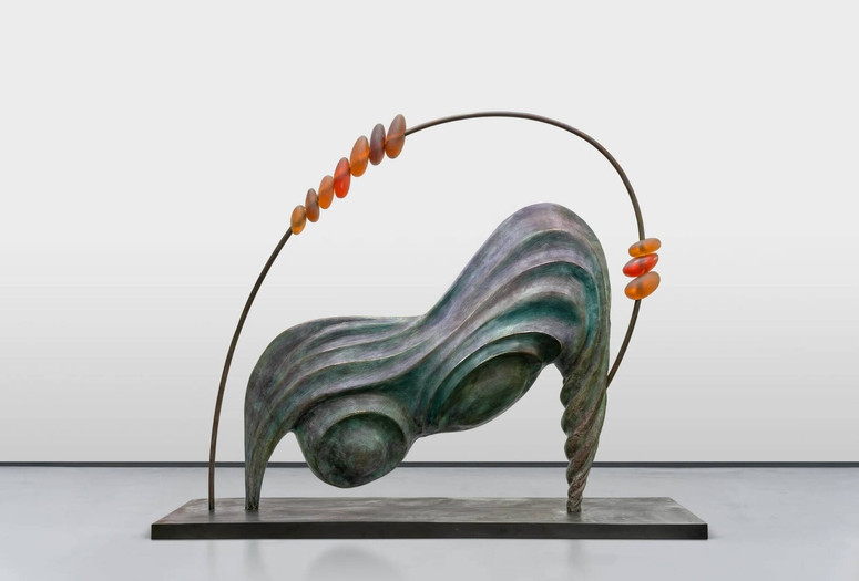 Camille Henrot. 73 / 37 (Abacus). 2024. Bronze, resin, rubber, and Japanese pigments. The Museum of Modern Art, New York. Gift of Sarah Arison Image description: A large bronze sculpture featuring smooth, flowing forms, like ripples in sand. It stands on two legs at each end atop a flat rectangular platform. Two round lumps sit on the underside of the sculpture. A thin metal arch circles above, with 10 large orange glass beads strung along it.