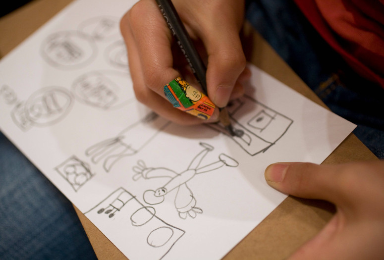 Close-up image of a child’s hands drawing a figure and everyday objects with a pencil on a white sheet of paper. Photo: On White Wall, Inc.