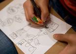 Close-up image of a child’s hands drawing a figure and everyday objects with a pencil on a white sheet of paper. Photo: On White Wall, Inc.