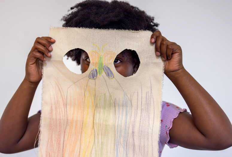 A child holds up a piece of canvas with a colorful drawing in front of their face with two holes cut into it. Photo: On White Wall, Inc.