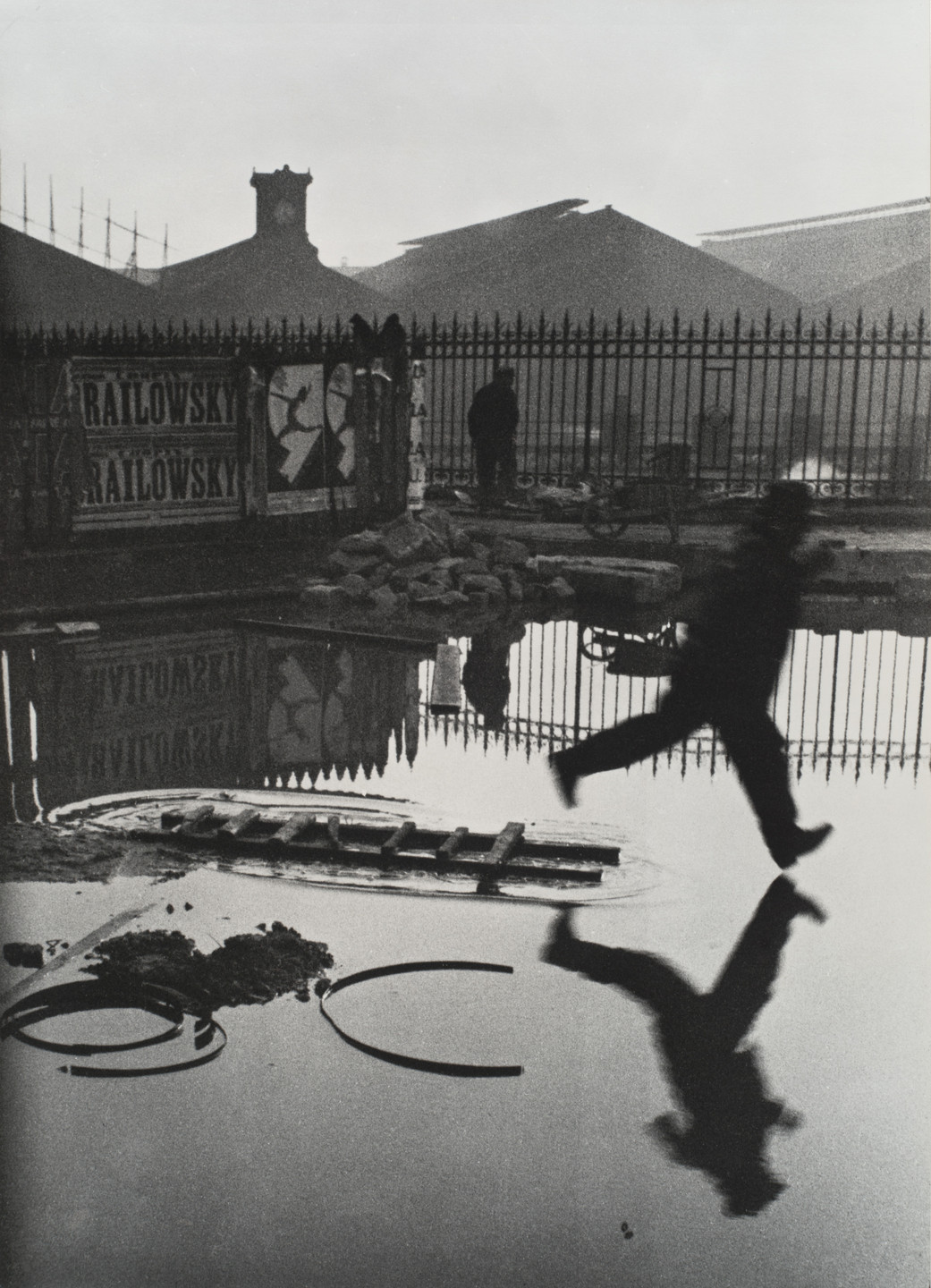 A person is captured in mid-air, jumping over a puddle with a ladder partially submerged in the water. The background includes a silhouette of buildings and a fence, under a gray sky. This is an AI generated description. To give feedback, email ai@moma.org.