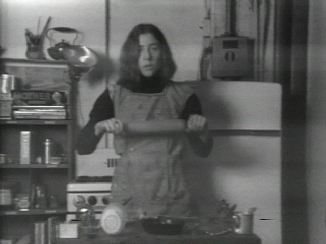 A person with shoulder-length hair, wearing an apron, stands in a kitchen holding a rolling pin. The black and white image shows kitchen shelves, a stove, and a refrigerator in the background. This is an AI generated description. To give feedback, email ai@moma.org.