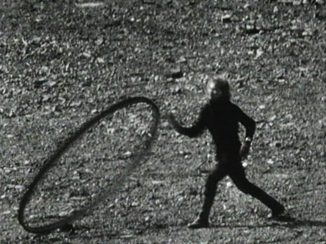 A person in dark clothing is standing on a rough, pebbled surface, rolling a large hoop. The image is in grayscale and predominantly features shades of black and white. This is an AI generated description. To give feedback, email ai@moma.org.