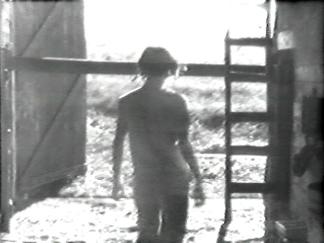 A person is seen from behind, walking towards an open door in a dimly lit area. The image is black and white, with a ladder propped against the wall to the right of the door. This is an AI generated description. To give feedback, email ai@moma.org.