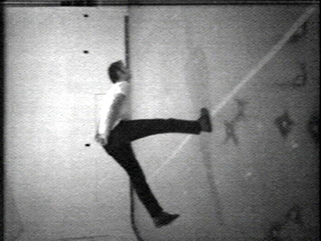 A grayscale image of a person in mid-air, leaning back with one leg extended upwards, and one leg bent downwards. Background appears to be a textured wall with some indistinguishable marks or shadows. This is an AI generated description. To give feedback, email ai@moma.org.