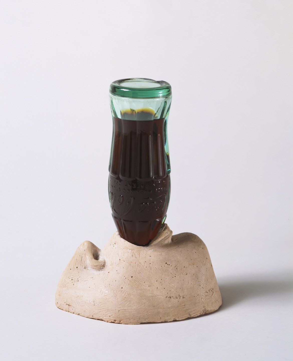 A green glass bottle filled with dark liquid is inserted upside down into a beige sculpture shaped like a nose resting on a flat surface. The background is plain white. This is an AI generated description. To give feedback, email ai@moma.org.