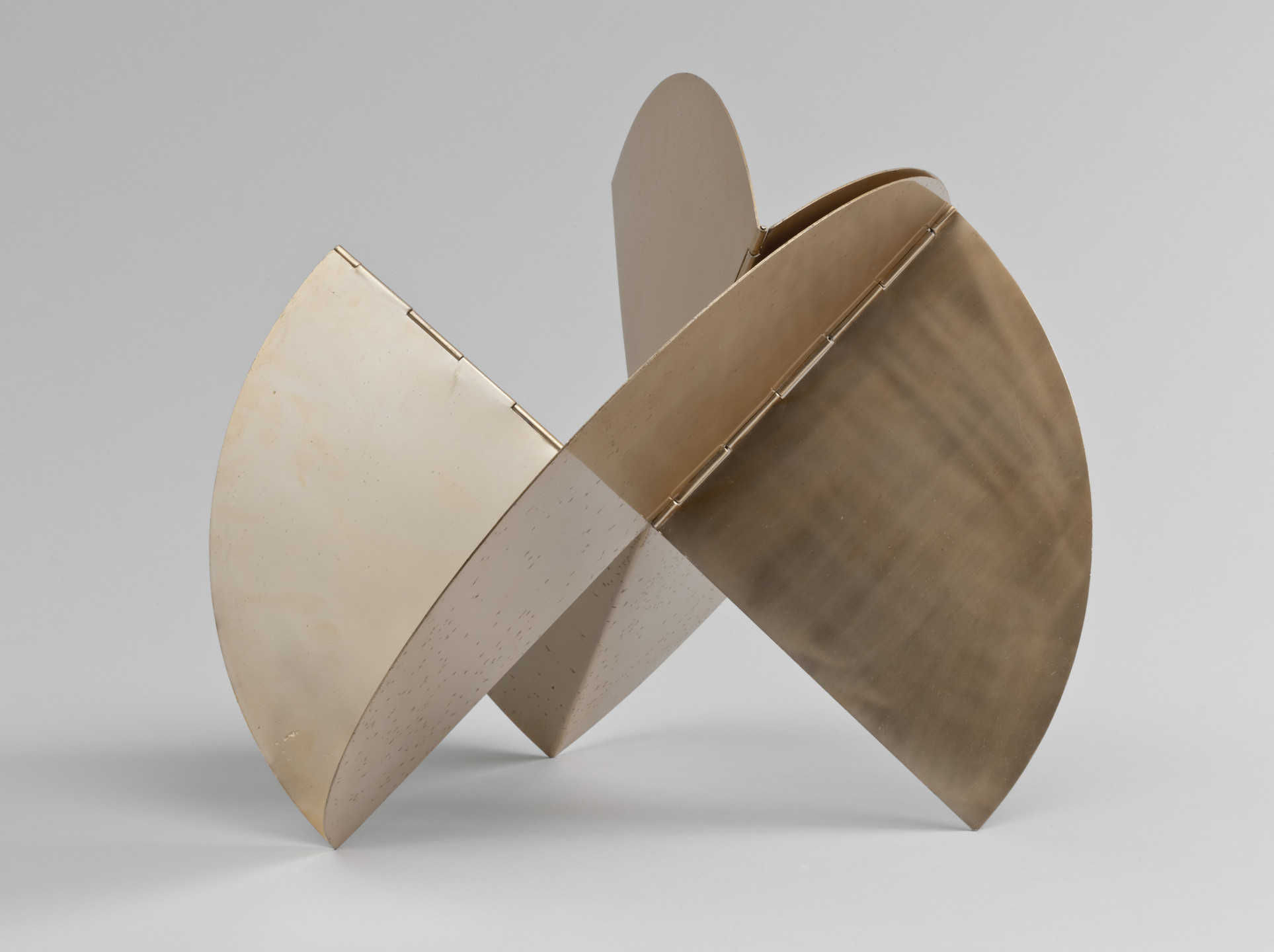 A sculpture composed of interconnected quarter-circle shapes in beige and brown hues, standing upright. The shapes intersect at their centers, forming an intricate, balanced structure. This is an AI generated description. To give feedback, email ai@moma.org.